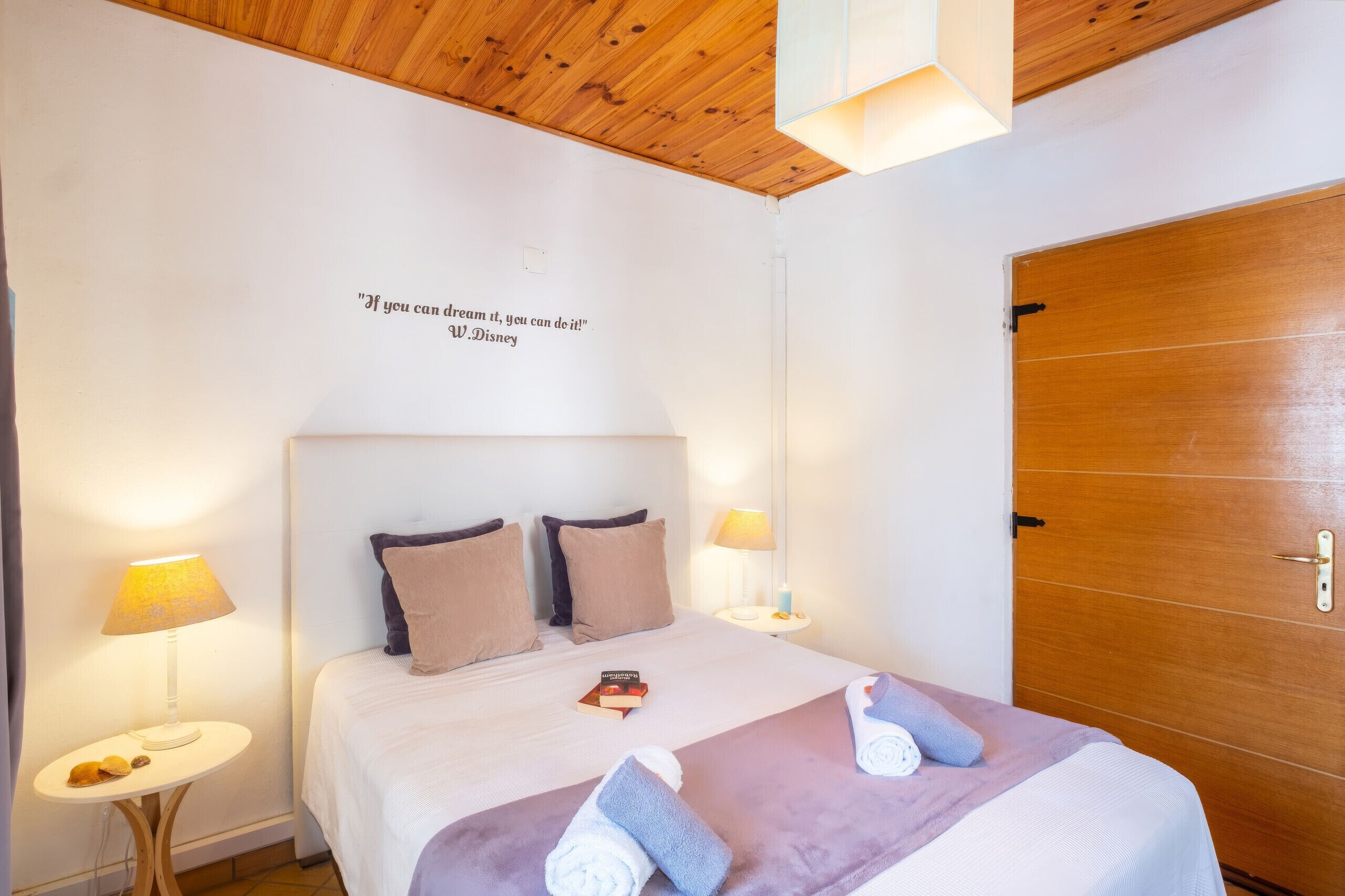 Une chambre d’hôtel avec un plafond en bois, un lit doté d’une tête de lit, deux lampes de chevet, une porte et une citation inspirante sur le mur.