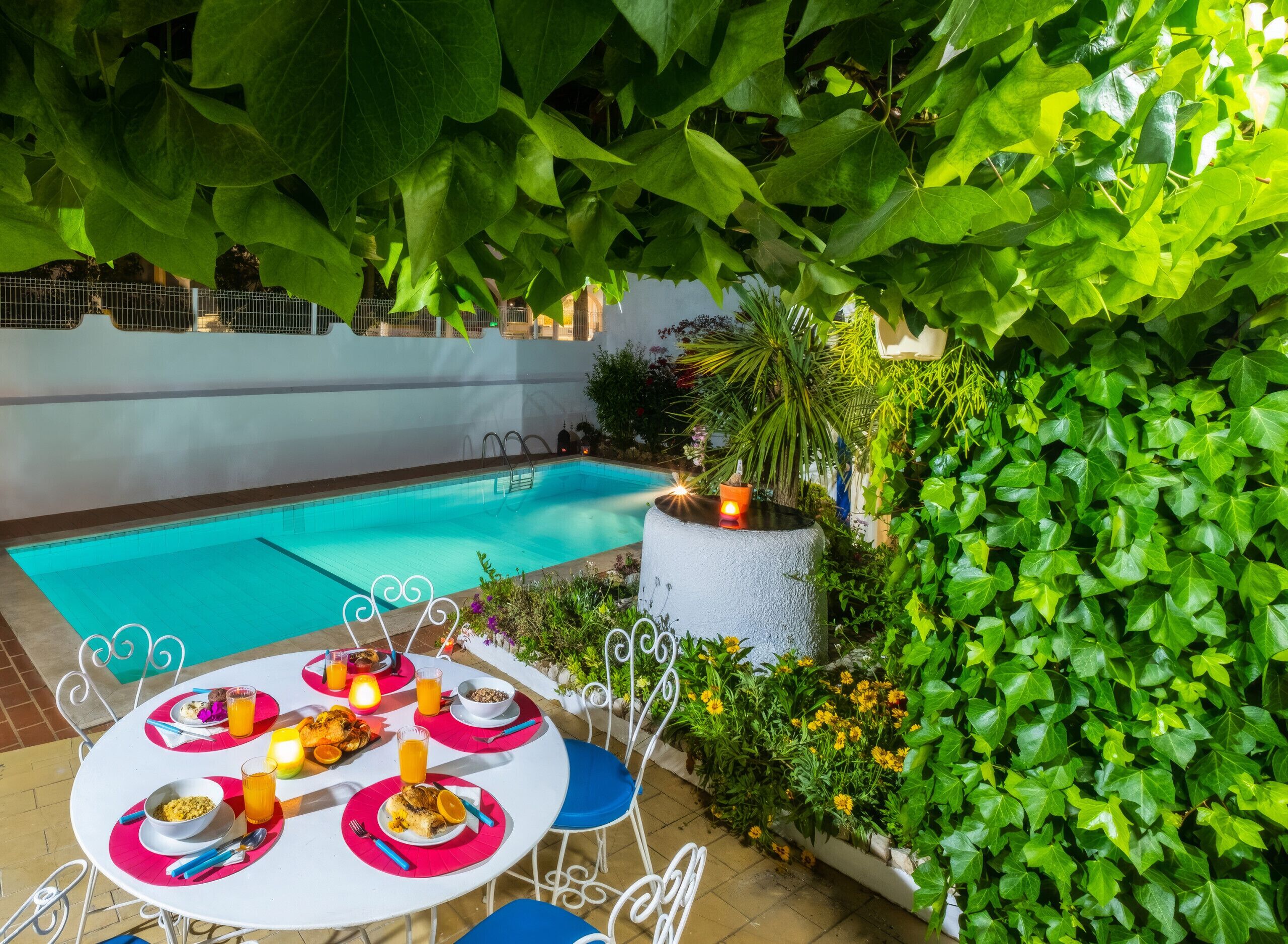 Ein Essbereich am Pool mit einem Tisch für zwei Personen, umgeben von üppiger Bepflanzung und einer weißen Wand.