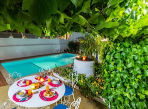 A poolside dining area with a table set for two, surrounded by lush greenery and a white wall.