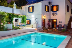 Uma área à beira da piscina com piscina transparente, mobiliário de exterior e um edifício branco com persianas azuis.