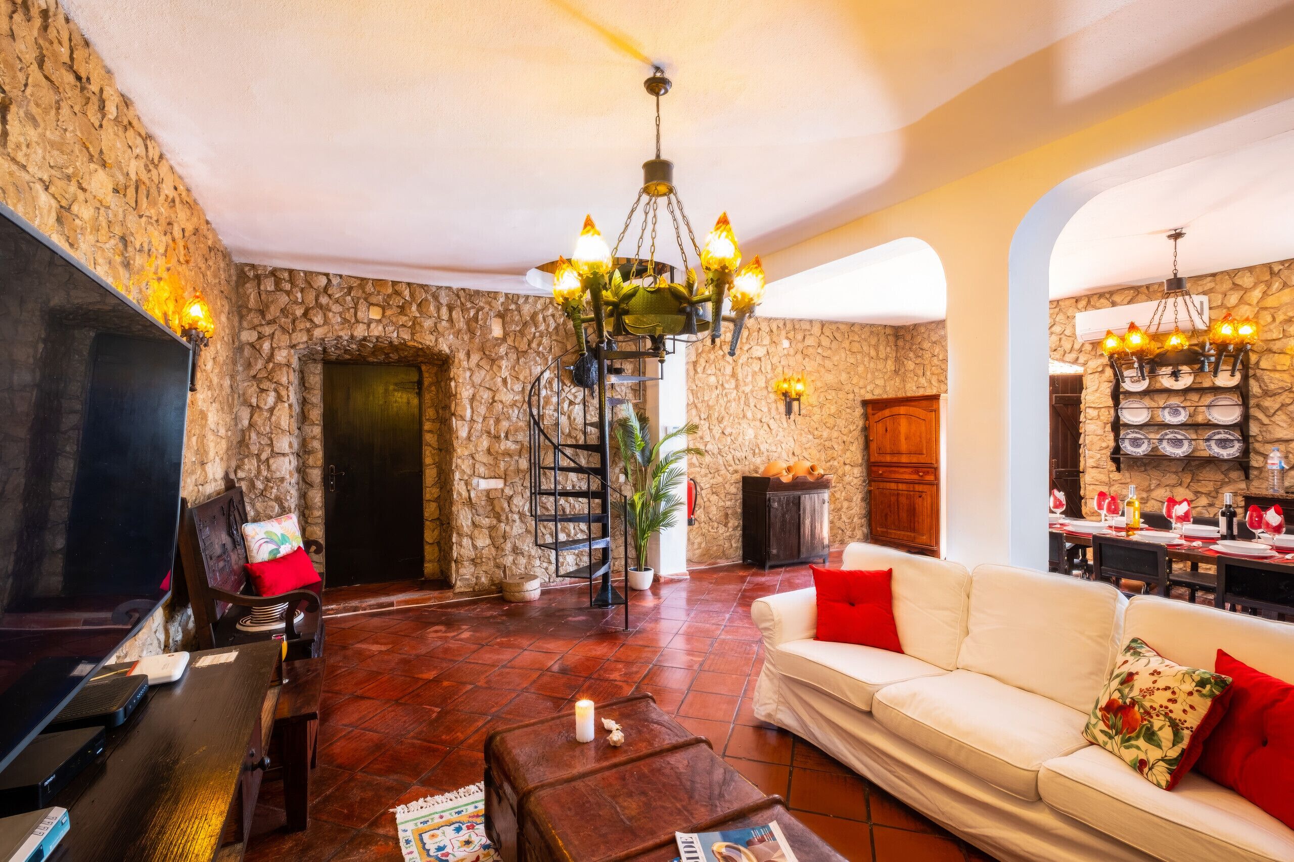 Un salon avec un mur en pierre, une cheminée, un canapé blanc, un téléviseur et un coin repas avec une table dressée pour quatre personnes.