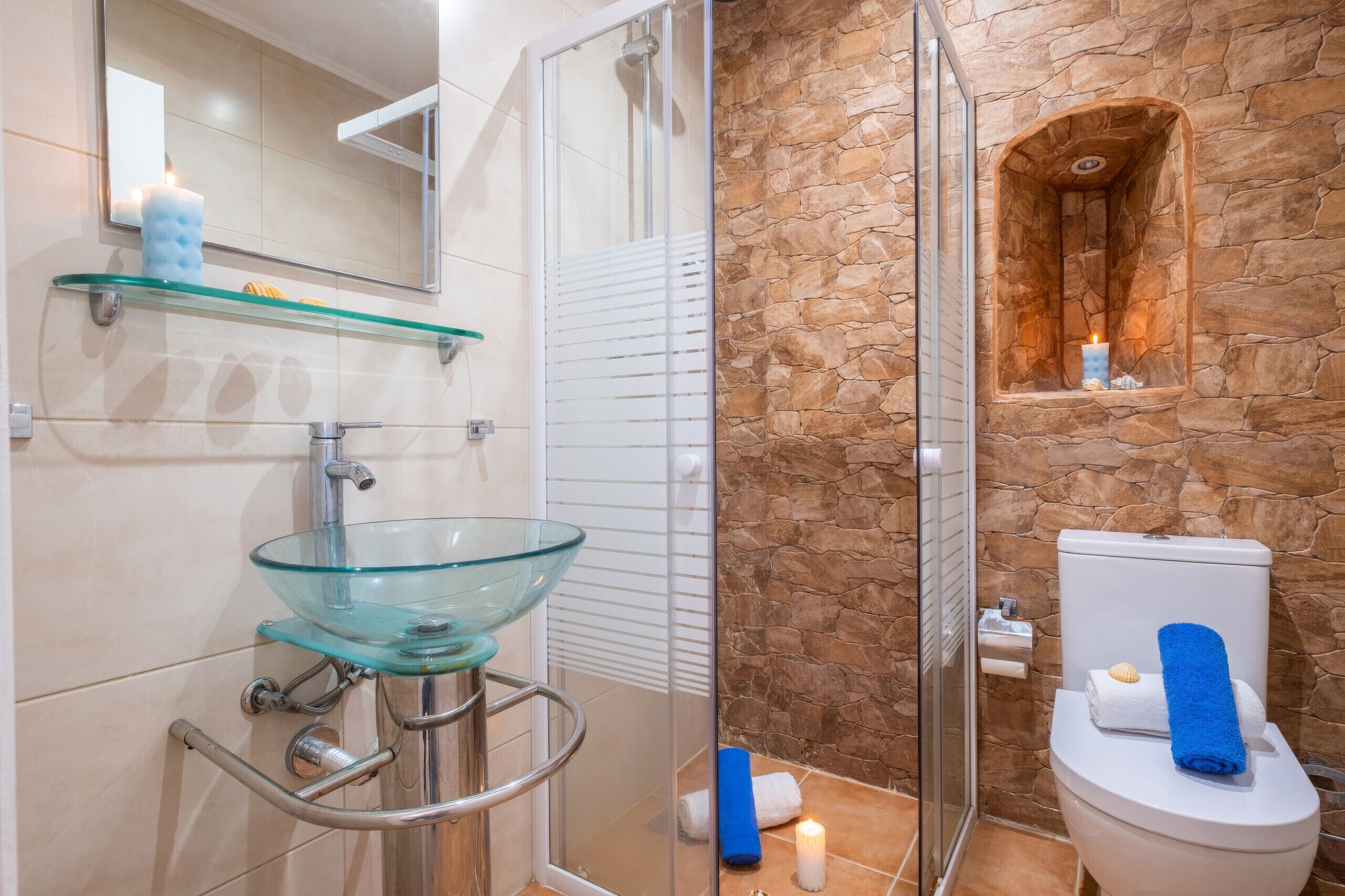 Un baño con lavamanos de vidrio, ducha, pared de piedra y un inodoro con toallas.