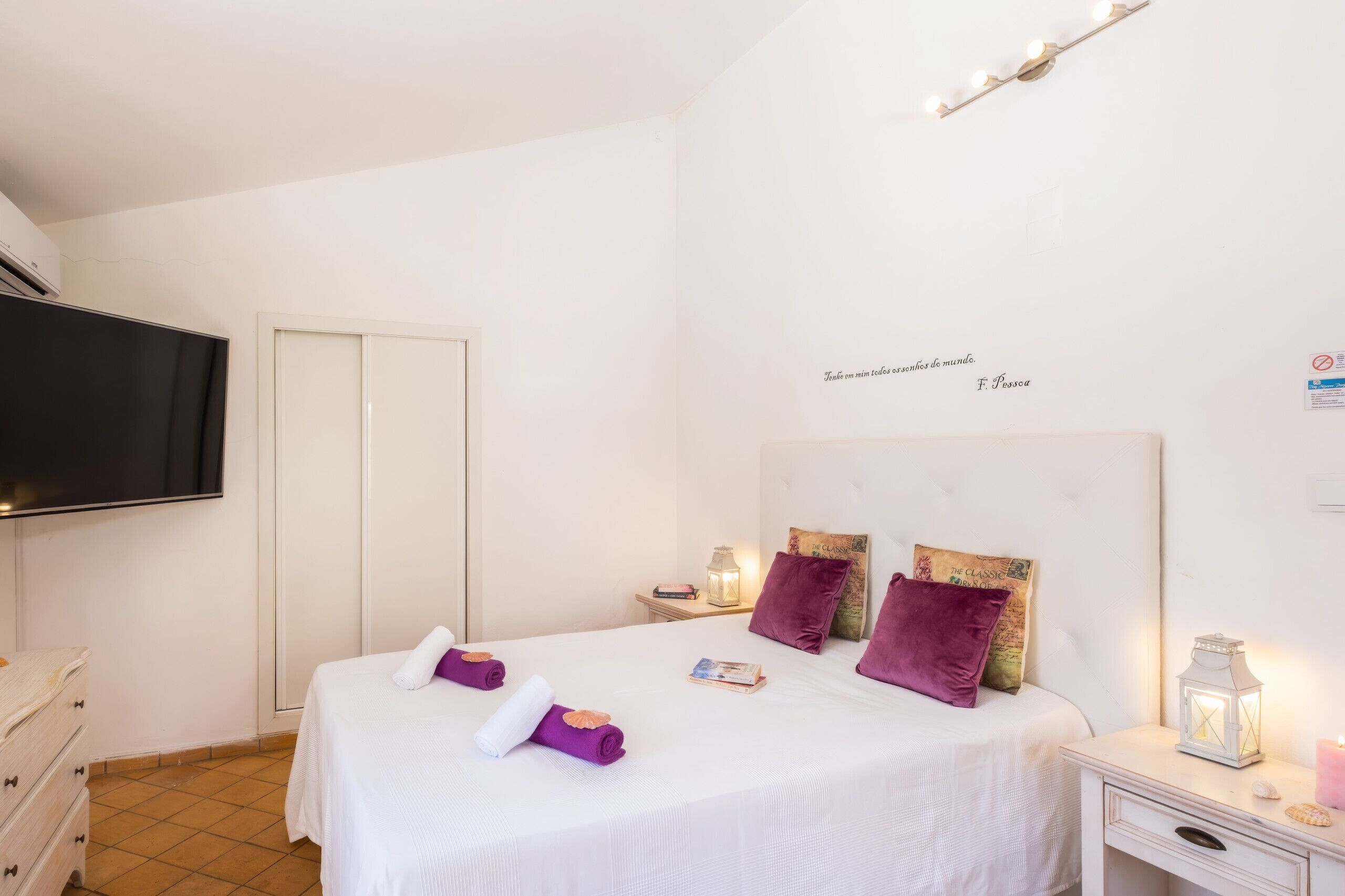 Quarto de hotel com cama, televisão, guarda-roupa e mesa de cabeceira com uma lâmpada e um livro.