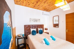 Hotellihuone, jossa on puinen katto, sängyssä on pääty, huoneessa on yöpöytä, seinälle kiinnitetty peili ja taulu, jossa on kuvattuna rantamaisema.