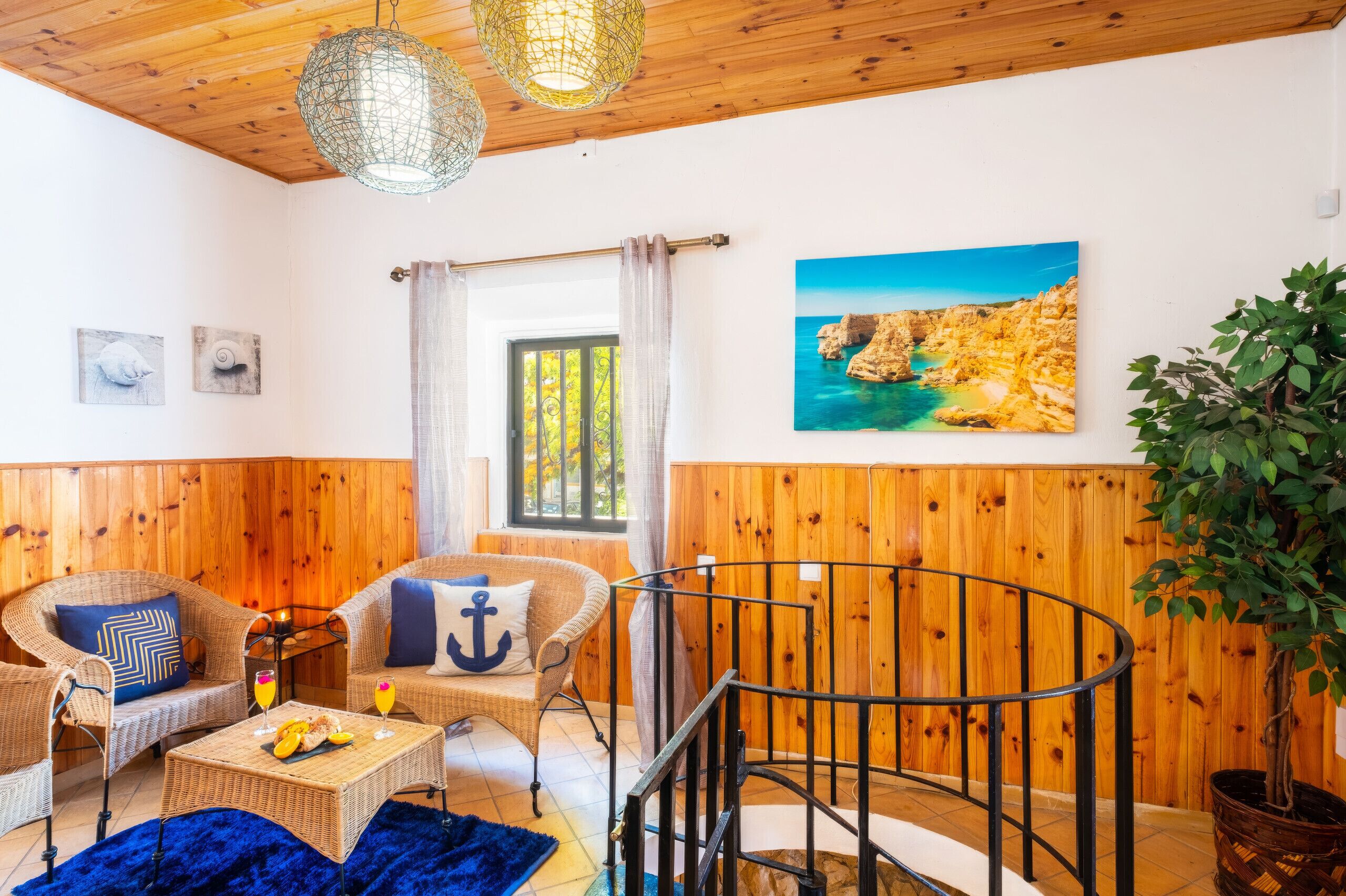 Una habitación con paneles de madera, un sofá de mimbre, una mesa de comedor con fruta, una escalera con barandilla y un cuadro de un paisaje costero.