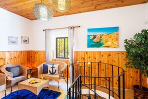 Ein Zimmer mit Holzwandverkleidung, einem Korbsessel, einem Esstisch mit Obst, einer Treppe mit Geländer und einem Gemälde von einer Küstenlandschaft.