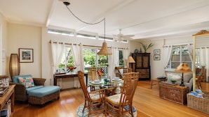 A spacious living room with a dining area, wooden flooring, wicker furniture, and large windows with curtains. - B.I. Plumeria Cottage -Summer Rates! Perfect Couples Getaway in Magical Puako (Kamuela)
