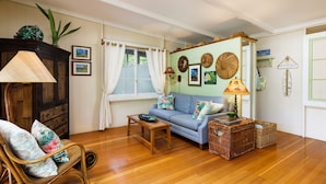 A living room with a wooden floor, a blue sofa, a wicker chair, and a wooden cabinet. - B.I. Plumeria Cottage -Summer Rates! Perfect Couples Getaway in Magical Puako (Kamuela)
