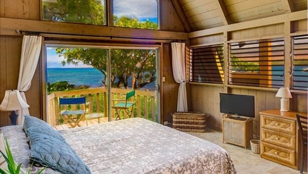 A bedroom with a large bed, a TV, a wooden dresser, and a balcony with a view of the ocean.