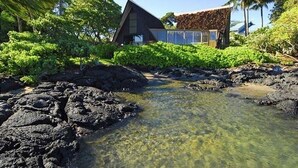 A house with a triangular roof, surrounded by lush greenery and a clear stream.