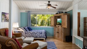 Un dormitorio con cama, televisor, ventilador de techo y ventana con vista a palmeras.