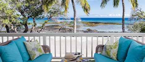 A balcony with wicker furniture, two drinks, and a hat on a table, overlooking a beach with palm trees and the ocean.