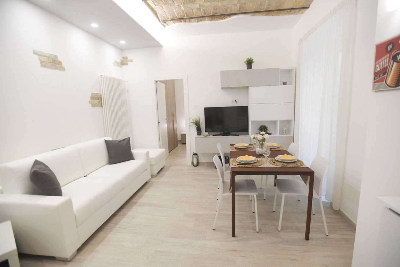 A modern living room with a white sofa, a dining table set for four, and a TV mounted above it.