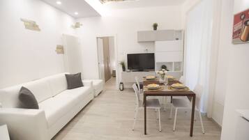 A modern living room with a white sofa, a dining table set for four, and a TV mounted above it.
