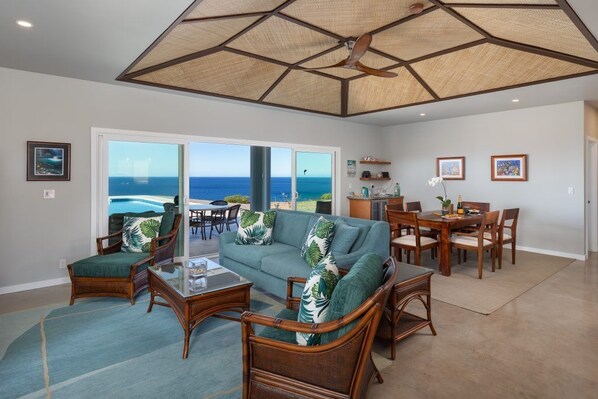 A spacious living room with a large window overlooking the ocean, a dining area with wooden furniture, and a ceiling fan. - B.I. Kohola Lani , Very private, Pool, Amazing Views! AC in bedrooms! (Kamuela)