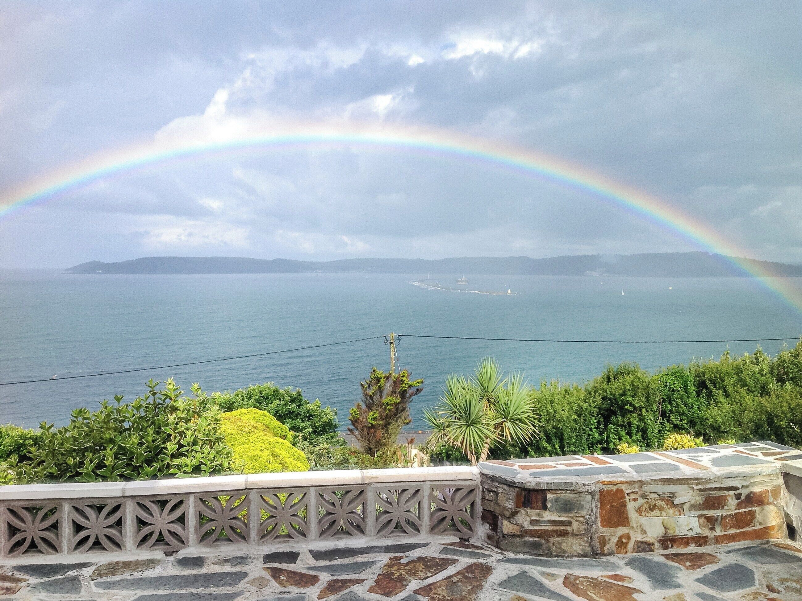 Vista sulla costa con un arcobaleno sul mare, un muro di pietra e vegetazione.