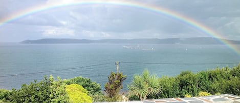 A coastal view with a rainbow over the sea, stone wall, and greenery.