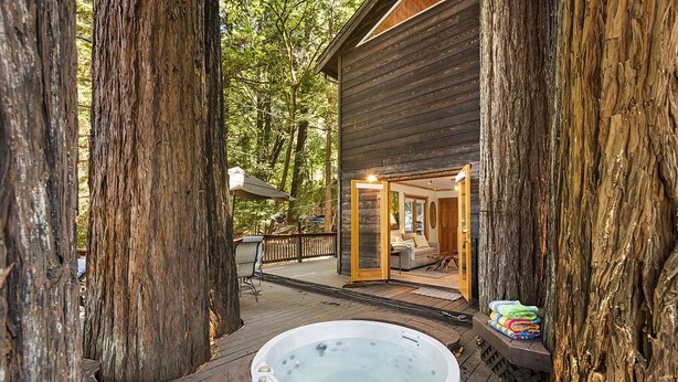 Une terrasse en bois avec un jacuzzi, entourée de grands arbres, qui mène à un chalet doté d’un porche et d’un coin repas.