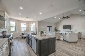 Uma cozinha espaçosa com uma ilha central, uma sala de jantar com mesa e cadeiras brancas, e uma sala de estar com sofá e televisão.