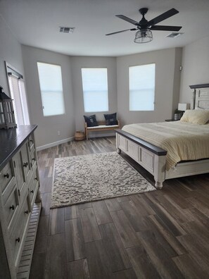 Quarto com cama, um banco, ventilador de teto e janelas amplas.