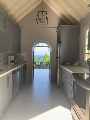 A modern kitchen with a view to a balcony, featuring stainless steel appliances, a hanging light fixture, and a tiled floor.