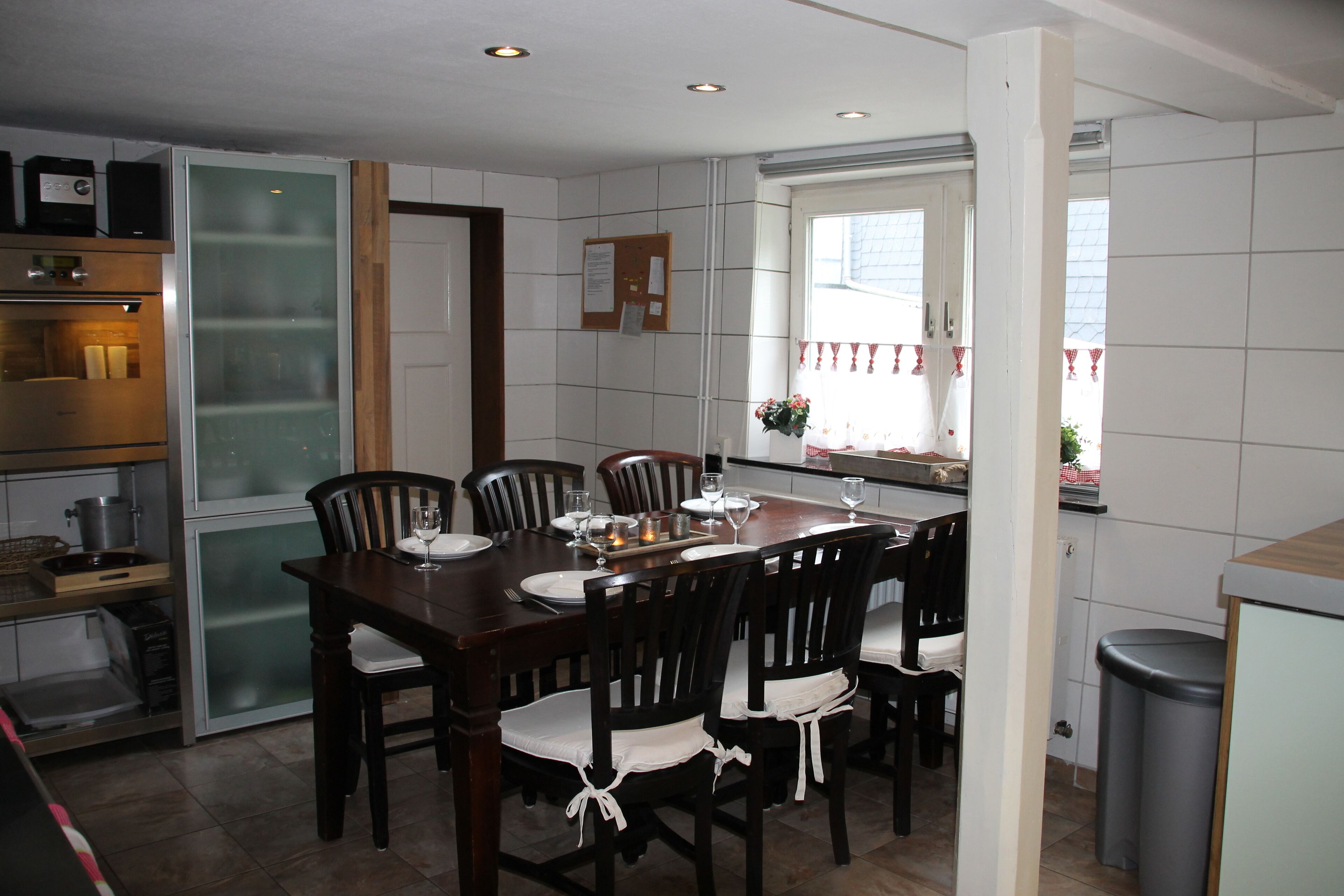 A dining area with a wooden table set for six, surrounded by chairs, a window with a view, and a kitchen area with appliances.