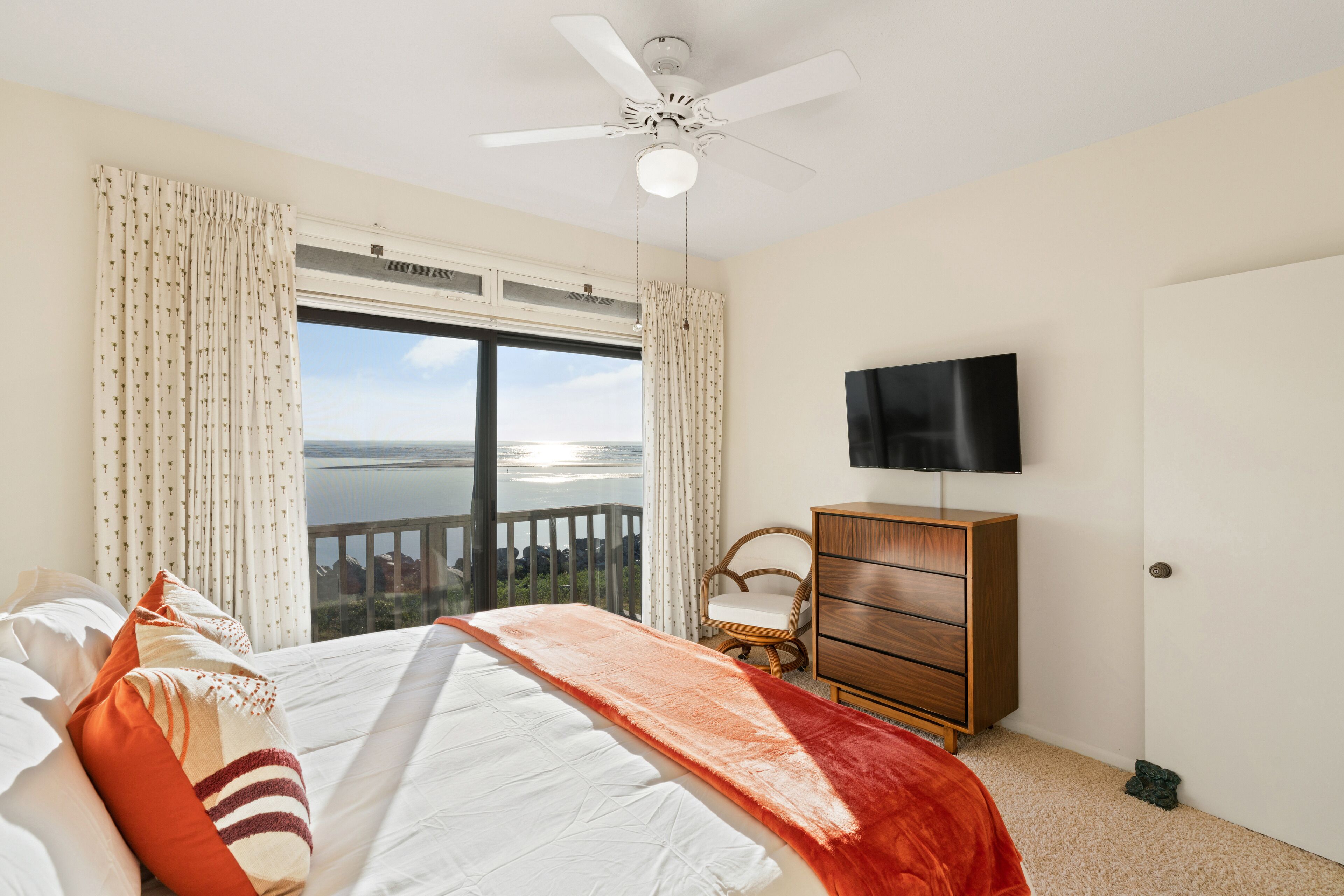Ein Schlafzimmer mit einem Bett, einer Holzkommode, einem Fernseher und einem Balkon mit Blick aufs Wasser.
