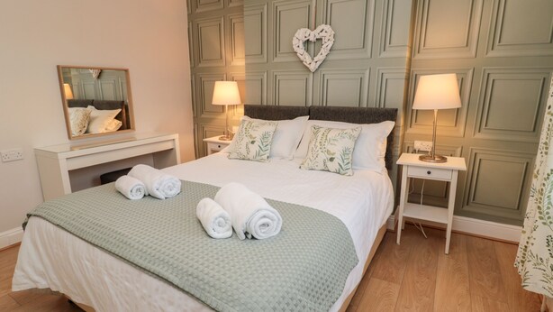 A neatly made bed with rolled towels, two bedside tables with lamps, a heart-shaped decoration on the wall, and a mirror above the bed. - Redshank (Morpeth)