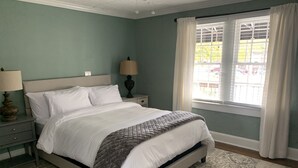 A neatly made bed in a room with a patterned rug, a nightstand with a lamp, and a window with curtains. - Cozy Cottage on Canton Street just steps away from nightlife, shopping and parks (Roswell)