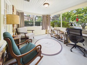 A room with a wooden rocking chair, a desk with a lamp, a modern office chair, and a view of greenery outside.
