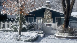 Een besneeuwde woonwijk met een stenen huis, een witte picket-hek en een met sneeuw bedekte boom.