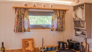 Una cocina con encimera de madera, un lavamanos, una ventana con cortinas florales y una estantería con platos y vasos.