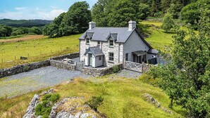 A stone house with a sloped roof, surrounded by a stone wall and a grassy field. - An impressive, detached stone-built cottage set within Gwydir Forest. (Betws-y-Coed)