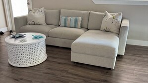 Een woonkamer met een beige hoekbank, een ronde salontafel en houten vloeren. Er hangen kunstwerken boven de bank en er is een raam aan de rechterkant.