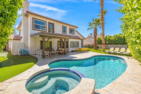 Une maison à deux étages avec une piscine dans le jardin, une terrasse dotée de mobilier d’extérieur et des palmiers.