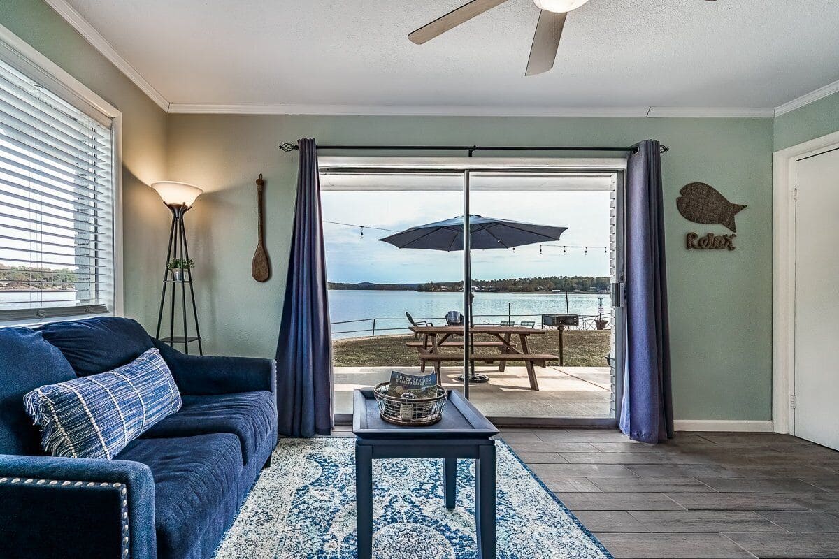 A living room with a dark blue sofa, a small table, and a view of a lakeside patio with a table and umbrella.