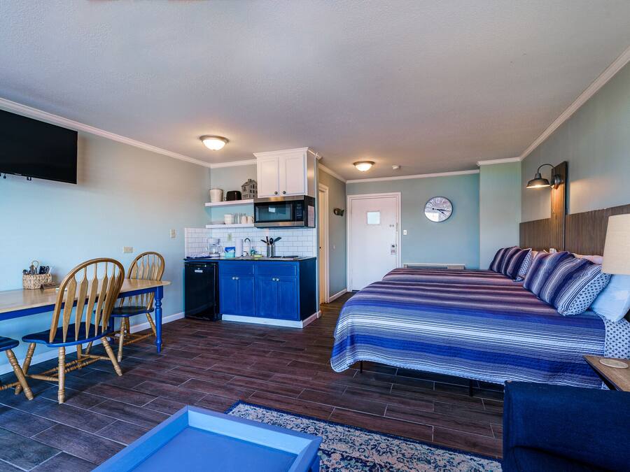 Una habitación compacta con cama, una zona de comedor con sillas, una kitchenette y un reloj en la pared.