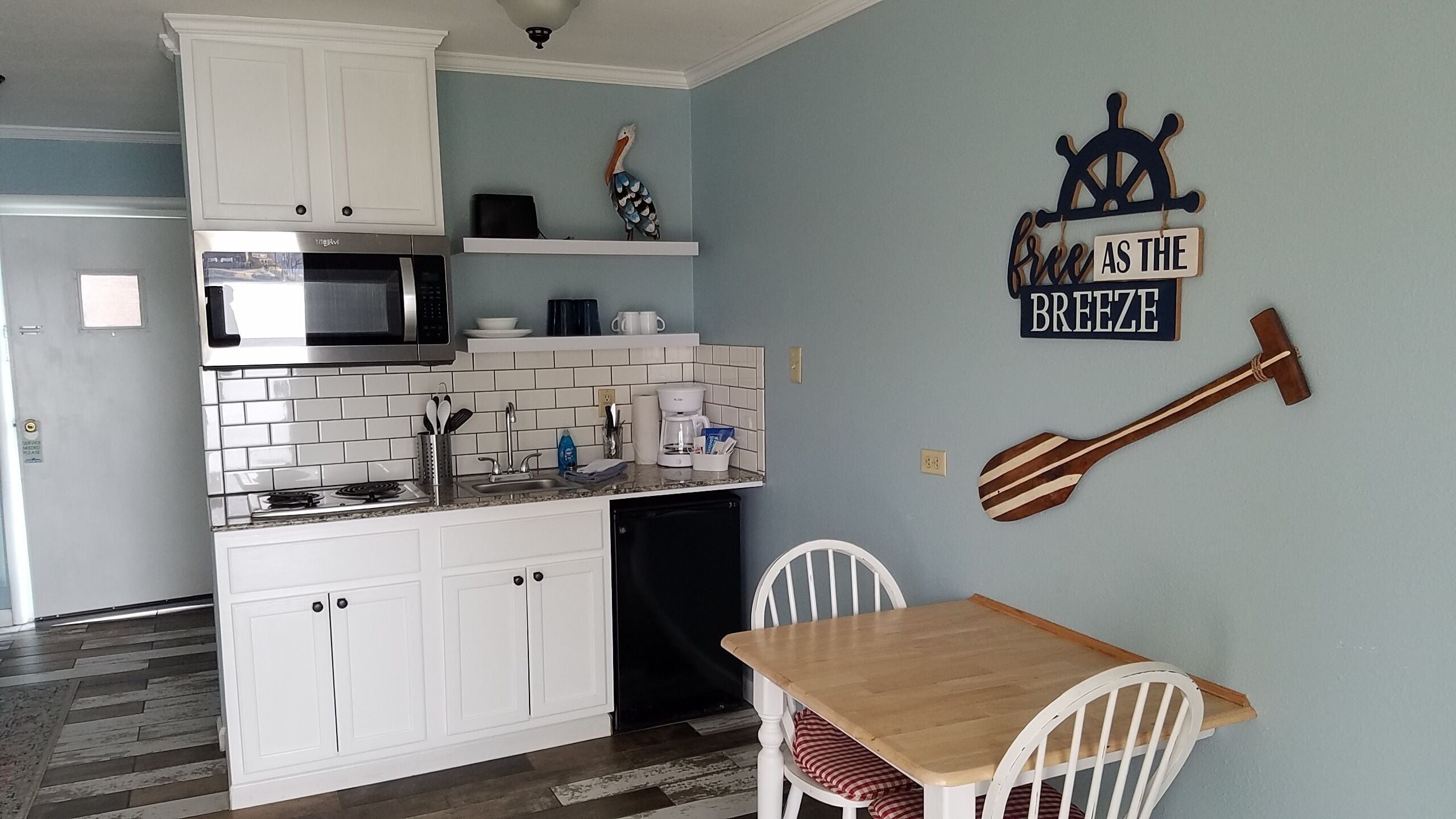 A compact kitchen with white cabinets, a microwave, and a coffee maker. There is a wooden table with chairs and a wall decoration of a ship's wheel and a sign that says "free as the breeze".