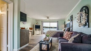 A spacious living room with a large window, a dark-colored sofa, a coffee table, and a television mounted on the wall. - Willow Beach Resort (Hot Springs)