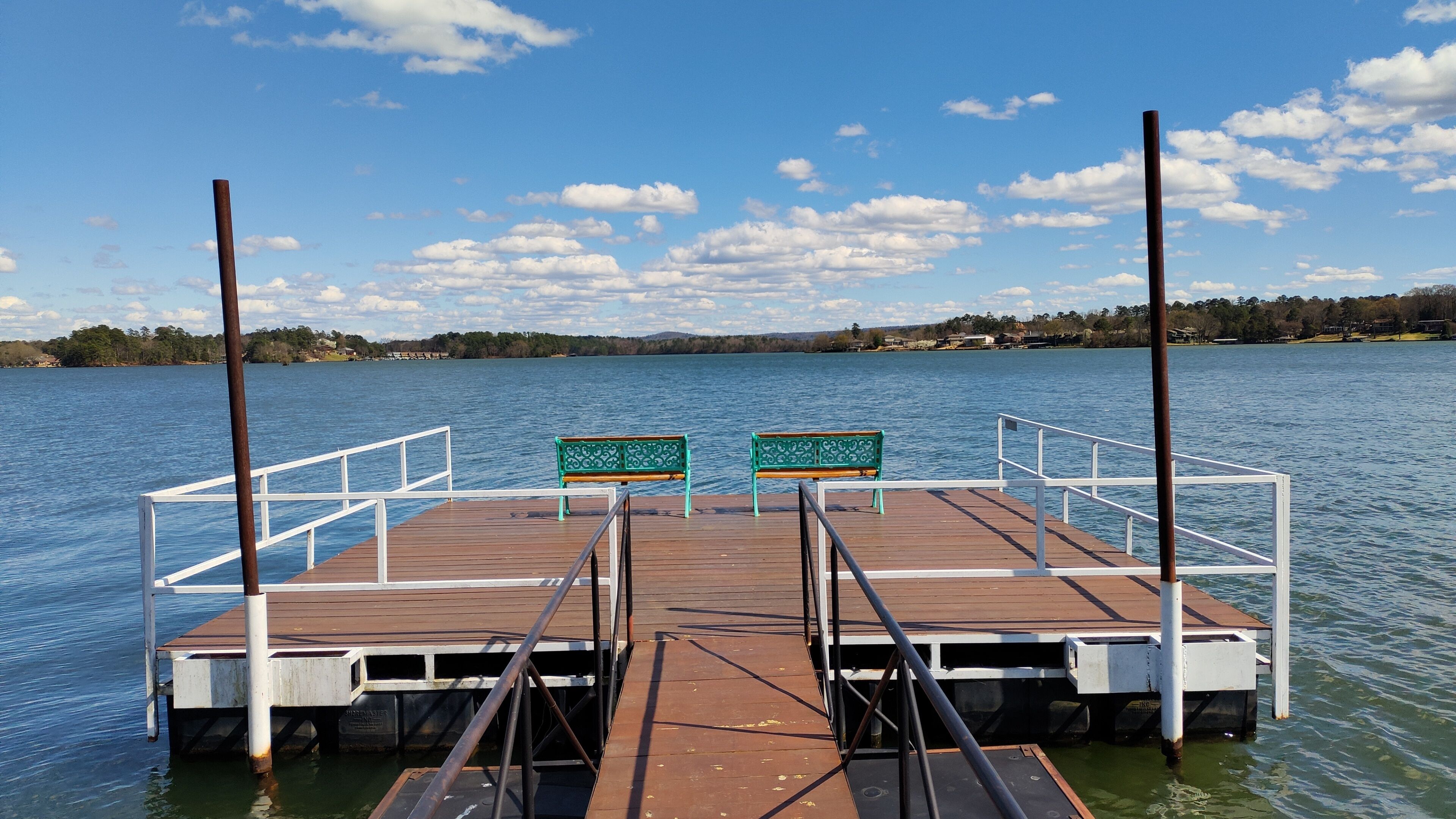 A wooden dock with two green benches and white railings, extending over calm water.