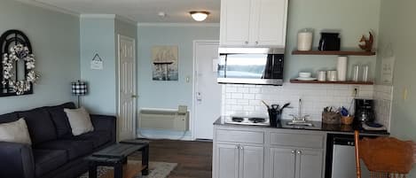 Una cocina compacta con microondas, estufa y lavaplatos. Hay una zona de comedor con una mesa y sillas. Un sofá negro con almohadones a rayas se encuentra en la sala.