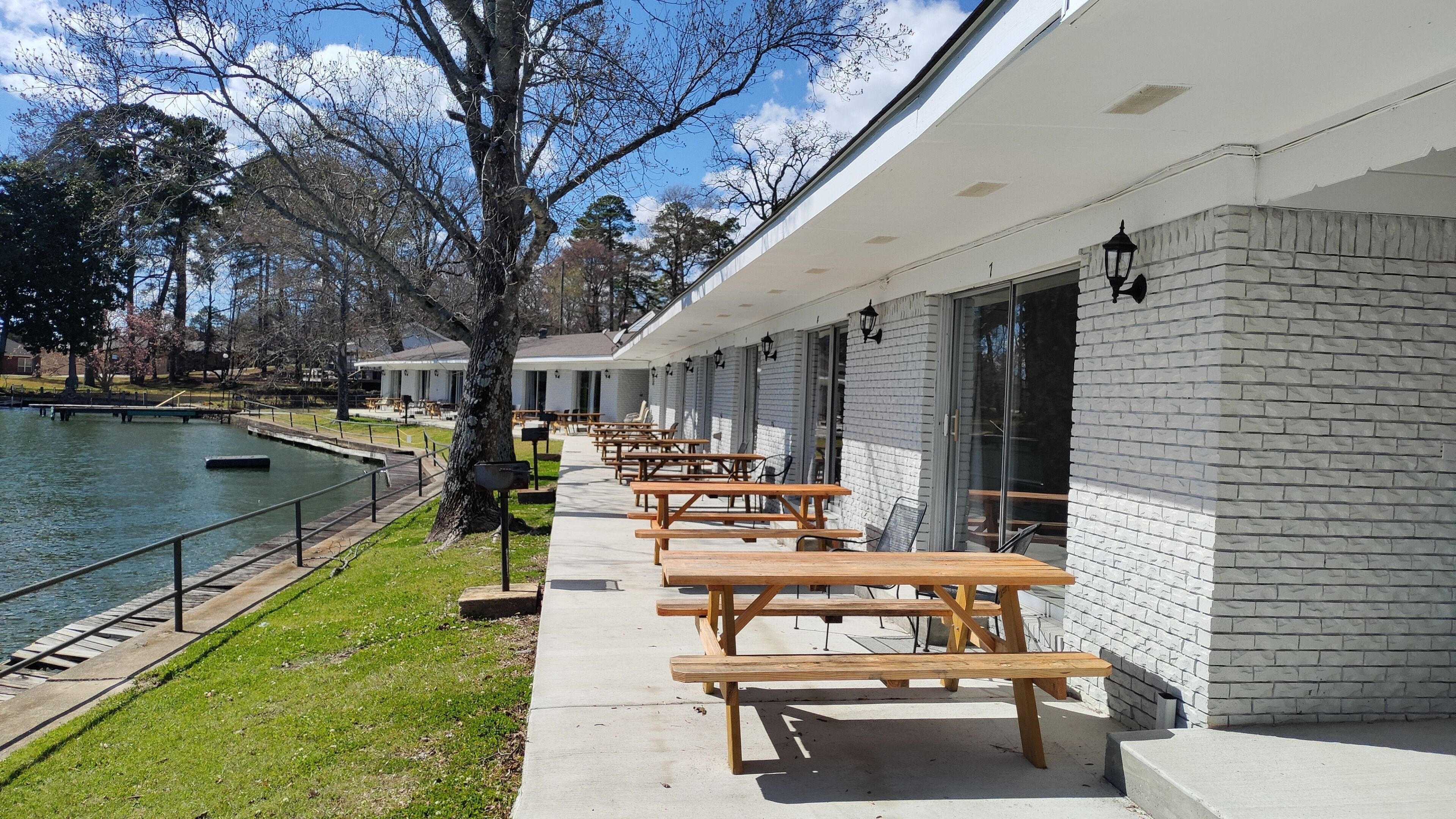 A row of wooden picnic tables outside a building with a brick facade, a pond, and trees in the background.