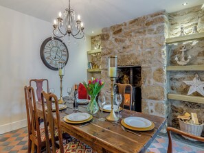A dining area with a wooden table set for four, surrounded by chairs. A chandelier hangs above, and a large wall clock is visible. A fireplace with a stone wall backdrop is in the background.