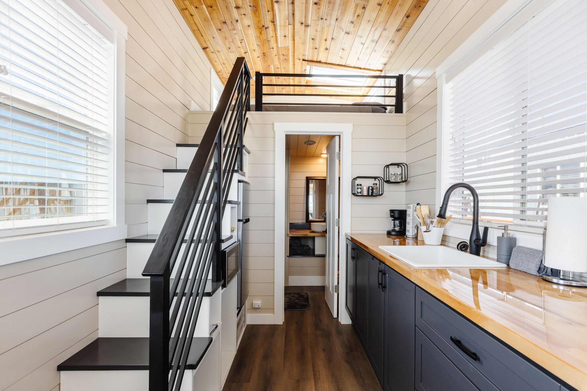 A modern kitchen with a wooden ceiling, a black staircase, and a view of the living area.