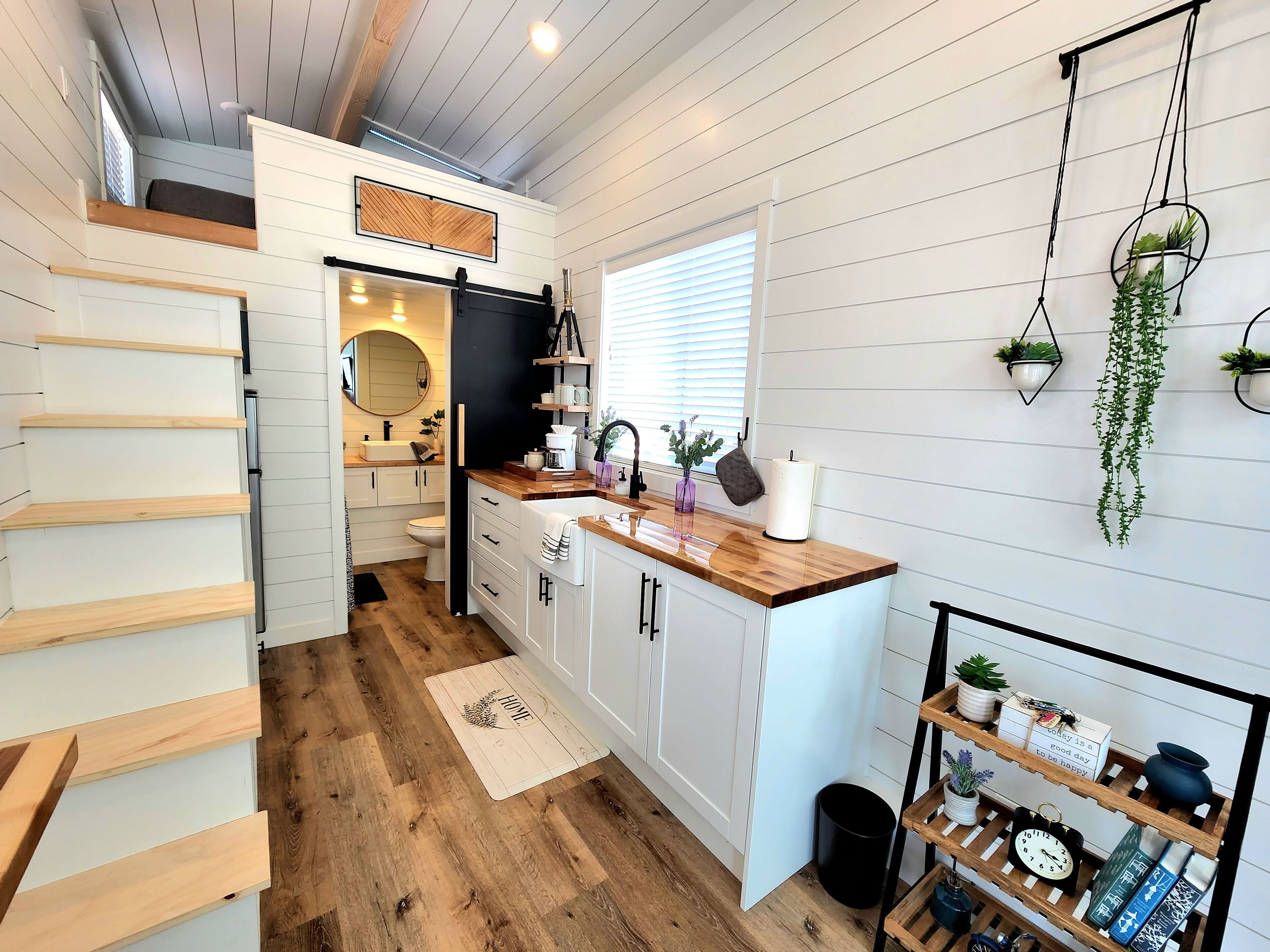 Een moderne keuken met witte kasten, een houten aanrecht en een zwart spatwand. Er is een raam met zonwering, een hangende plant en een plank met keukengerei.