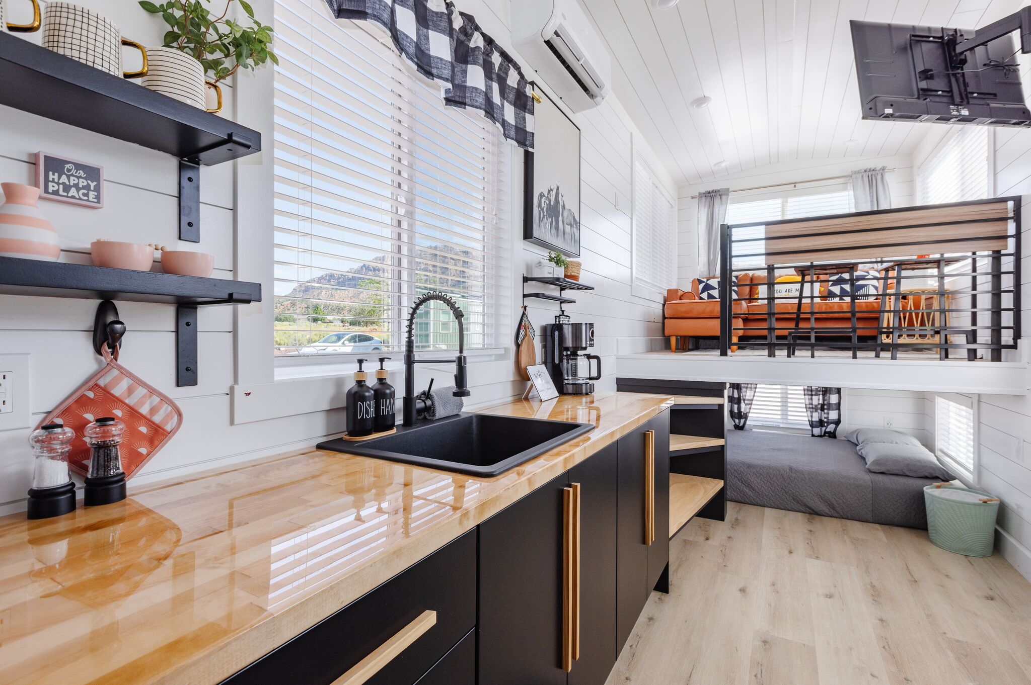 A modern kitchen with a wooden countertop, black cabinets, and a built-in oven. There is a window with blinds, a sink, and a coffee machine.