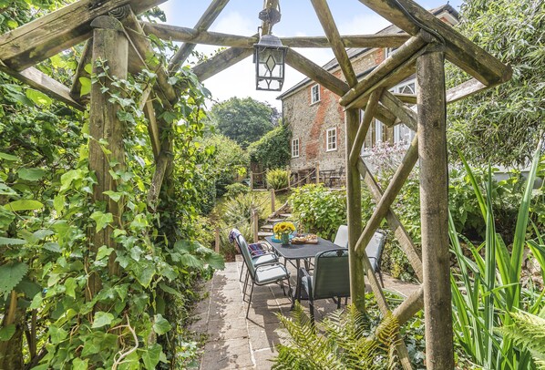 Une terrasse de jardin avec une table et des chaises, entourée d’une végétation luxuriante et bordée d’un chemin en pierre.