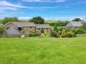 A stone building with a sloped roof, surrounded by a grassy field and trees. - Twinkles Cottage-UK12900 (Pelynt, near Looe)