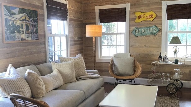 A cozy living room with a sofa, two wicker chairs, a coffee table, and a ceiling fan. There are two windows with blinds, a lamp, and a sign indicating '90 miles to Cuba'. - Old Town Classic with plenty of charm, porch swing and parking (Key West)