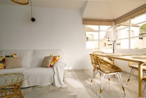 Una habitación con un sofá blanco, sillas de mimbre, una mesa de vidrio y un ventanal con vistas a edificios.